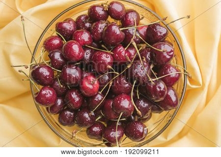 Ripe bright cherry with droplets of water in a transparent bowl on a yellow tablecloth