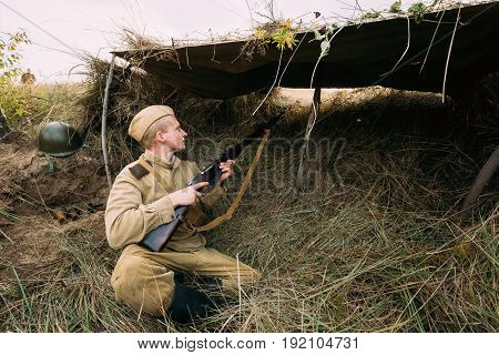 Dyatlovichi, Belarus - October 1, 2016: Reenactor Dressed As Russian Soviet Red Army Soldier Of World War II Hidden Sitting And Aiming Rifle Weapon From Trench, Hovel, Ambush In Autumn Meadow