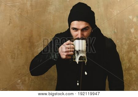 man or bearded hipster with long beard and moustache drinking beer with foam from glass mug in black hat and jacket on beige wall. Alcohol bad habits addictive lifestyle refresher convive