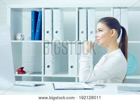 Pensive young business woman sitting turning sideways at the dask with desktop monitor and looking at the window.