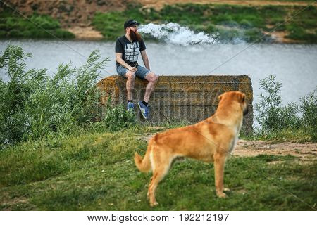 Vape Bearded Man In Real Life. Portrait Of Young Guy With Large Beard In A Cap And Sunglasses Having
