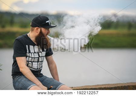 Vape Bearded Man In Real Life. Portrait Of Young Guy With Large Beard In A Cap And Sunglasses Having