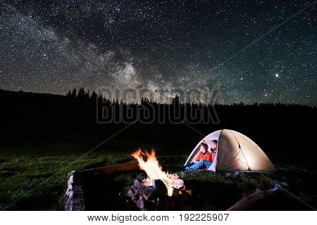Night Camping In The Mountains. Romantic Couple Tourists Sitting In The Illuminated Tent Near Campfi