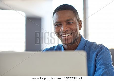 Portrait of a smiling young African businessman sitting confidently at his desk in a large modern office working on a laptop