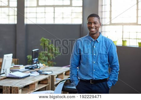 Portrait of a confident young African businessman smiling while standing with his hands in his pockets in a large modern office