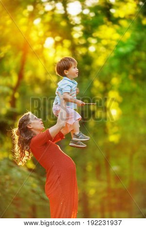 A pregnant woman in a red knitted dress high raises her son standing on the edge of the forest