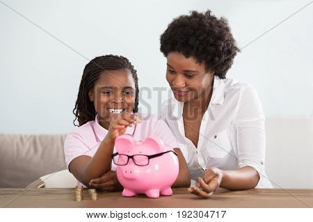 An African Girl Sitting With Her Mother Inserting Coins In Pink Piggy Bank At Home