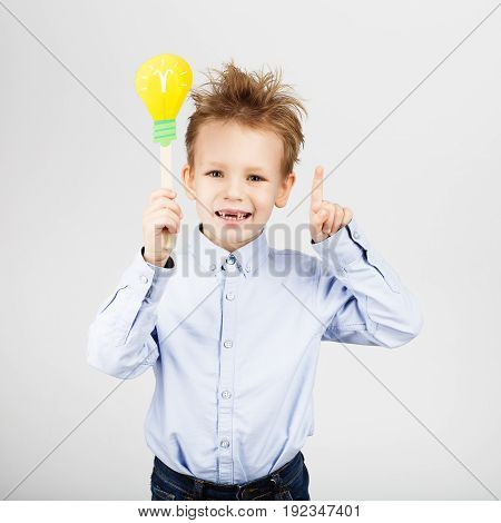 Cute Little School Boy With Yellow Paper Lightbulb Against A White Background. Cheerful Smiling Kid