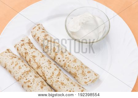 Three pancakes roll with stuffing and sour cream in a bowl in a white plate on a delicate peach-orange background close-up