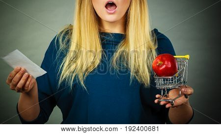 Shocked woman holding shopping basket with fruits looking at bill receipt being scared of huge prices