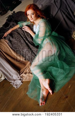 Tender redhead woman in transparent mint peignoir and white lingerie sitting on the floor near her bed in bedroom waiting for a man