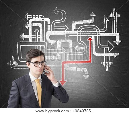 Young businessman wearing glasses a suit and a yellow tie. He is talking on his smartphone. Blackborad background with arrow labyrinth