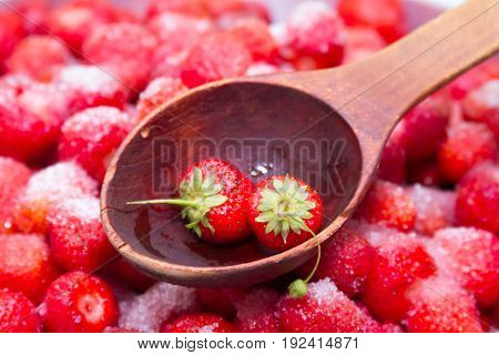 Against a background of strawberries with sugar a wooden spoon for jam on which lies a few strawberries with green leaves.