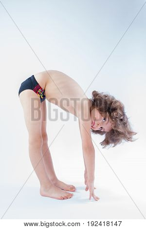 Curly boy in swimming trunks stands sideways making lean forward looking at the camera. Blue background.