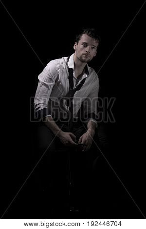 Portrait of young, attractive man in unbottoned, creased white shirt with upturned sleeves and loose tie sitting in the dark