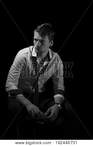 Black and white portrait of young, attractive man in unbottoned, creased white shirt with upturned sleeves and loose tie sitting in the dark