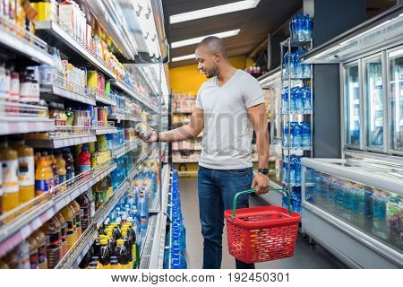 African man shopping in beverage section at supermarket. Black man doing shopping at market while buying cold drink. Handsome guy holding shopping basket reading nutritional values of product.