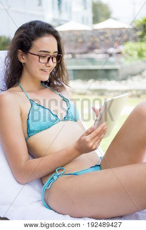 Portrait of young Caucasian woman wearing bikini and glasses sitting in chaise lounge and reading e-book or networking on digital tablet during vacations