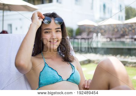 Portrait of young Caucasian woman wearing bikini and sunglasses sitting in chase lounge at hotel, sunbathing, looking at camera and smiling