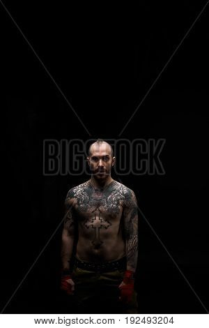 Portrait of young Caucasian male with tattoos all over his chest and arms standing in dark studio staring at camera having angry mean and aggressive look demonstrating his readiness for fight