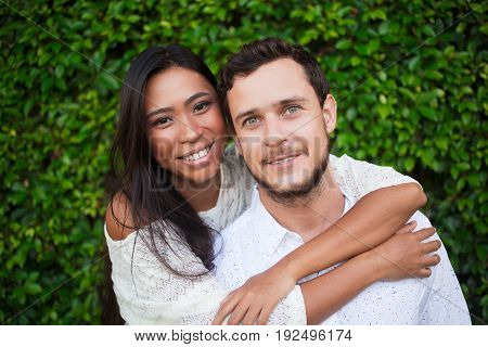 Closeup of affectionate young Asian woman embracing Caucasian boyfriend from behind with green leaves wall in background. They are looking at camera. Front view.