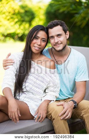 Closeup portrait of smiling young Caucasian man embracing Asian girlfriend. They are looking at camera and sitting on chaise longue with blurred trees in background. Front view.