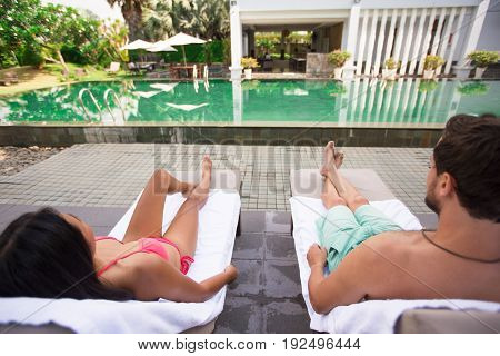 Closeup of young couple lying on chaise longues near swimming pool. They are back to camera.