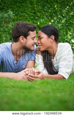 Closeup portrait of smiling young multi-ethnic couple holding hands, touching noses with their eyes closed and lying on patio grass with green leaves wall in background. Front view.