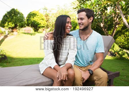 Closeup portrait of smiling young multi-ethnic couple embracing tenderly and sitting on chaise longue with blurred plants in background