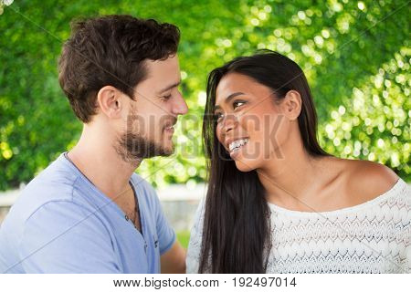 Closeup of smiling young interracial couple looking at each other with blurred green leaves wall in background