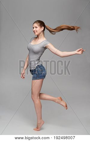 Beautiful barefooted full length female in denim shorts and grey tshirt jumping over grey background with hair ponytail flying in air