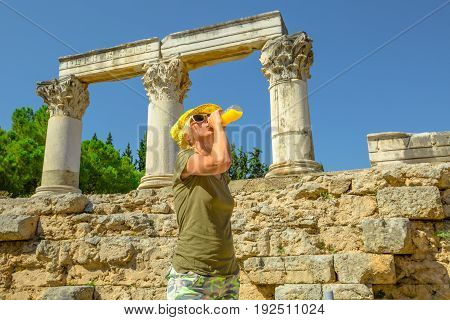 Young happy tourist drinks orange juice at Archaeological Site of Ancient Corinth in Greece. Young woman holding refreshing drink while visiting Temple of Ottavia in hot day. Tourism travel concept.
