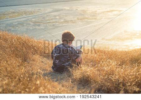 Boy reading book in yellow grass high in the hills, watching beautiful sunset above Bay Area, rear view