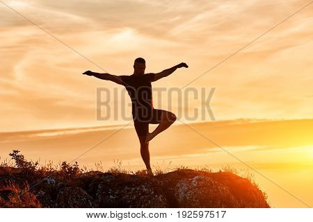 Silhouette of the man practicing yoga on the hill against beautiful sunset. Doing yoga exercises on the nature. Horizontal photo of young sportsman. Beautiful landscape and sky with clouds. Concept of the healthy lifestyle.