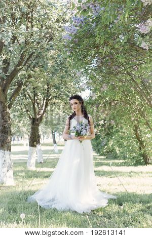 Tender beautiful cute girl bride in a white air dress with a bouquet of lilacs in her hands walking through the park on a sunny spring day. Photo in gentle colors