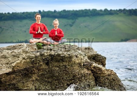 Sporty mother and daughter doing yoga on the rock near beautiful river. Beautiful mother and daughter dressed in the sportwear and barefoot. Horizontal photo with beauty landscape. Concept of the healthy and active lifestyle.