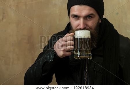 hipster with long beard and moustache drinking beer with foam from glass mug in black hat and jacket on beige wall. Alcohol bad habits addictive lifestyle refresher convive