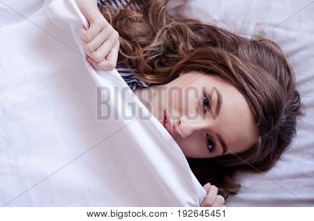 Beautiful young woman lying down in the bed after sleeping. Teen girl with open eyes covers her face with white blanket. Morning concept. View from above with place for your text. Copy space.
