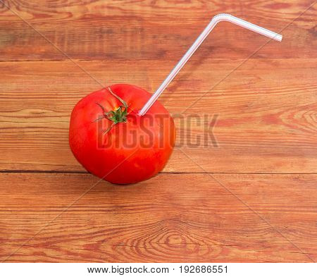 Big fresh ripe tomato with bendable drinking straw inserted into it on a surface of the old wooden planks