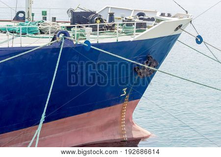 Bow of the old cargo ship with a blue starboard moored by several mooring ropes to the berth of the sea cargo port closeup