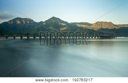 Sunrise lights the dawn sky above Hanalei Bay and pier with the Na Pali coast in the background near Hanalei, Kauai, Hawaii