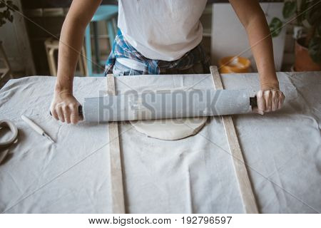 Artist rolls raw clay using big grey rolling pin on table covered with industrial fabric in workshop
