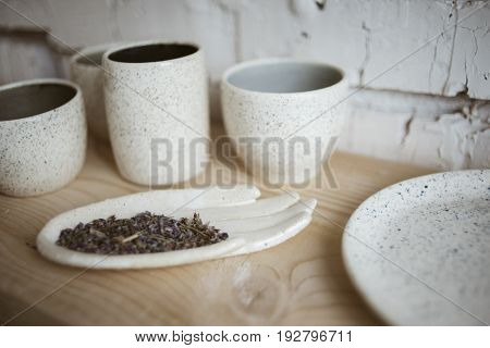 Stylish chamotte ceramic glazed hand with dry lavender and handmade cups on wooden shelf with white brick wall on background in workshop
