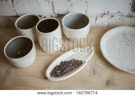 Stylish chamotte ceramic hand with dry lavender and handmade cups on wooden shelf with white brick wall on background in workshop