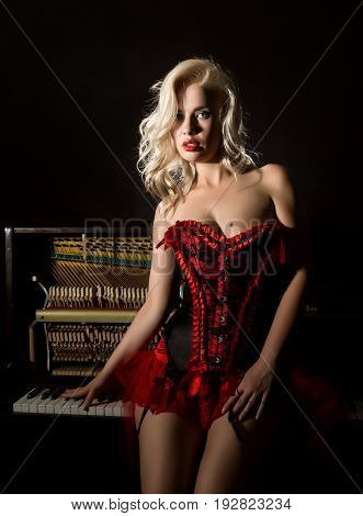 Young beautiful girl in red corset standing near the piano. Vintage style beautiful woman. Old fashioned makeup and retro finger wave hairstyle.