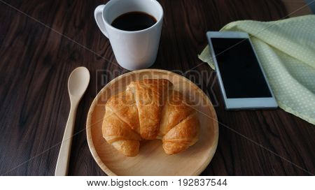 Croissant  on wooden plate with spoon, coffee cup and mobile phone on table