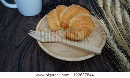 Croissant  on wooden plate with fork, coffee cup and wheat on table