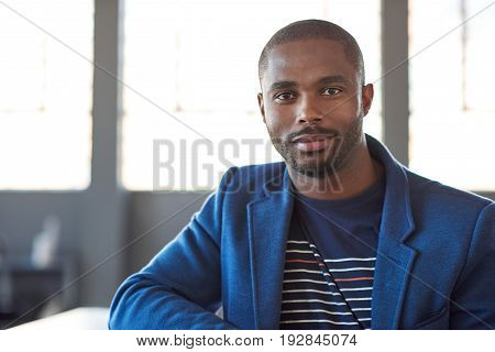 Portrait of a confident young African businessman in a blazer smiling while standing alone in a large modern office