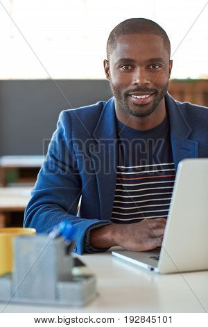 Portrait of a confident young African businessman smiling while sitting at his desk in a large modern office working on a laptop