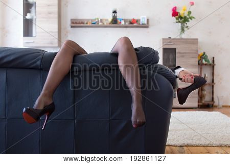 Businesswoman hanging sexy long legs at home on sofa after workday holding high heels